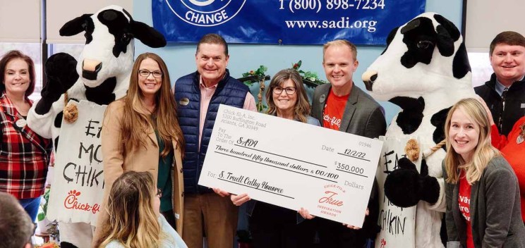 A group of people and the Chick-fil-A cow mascot celebrate indoors while holding a large check.