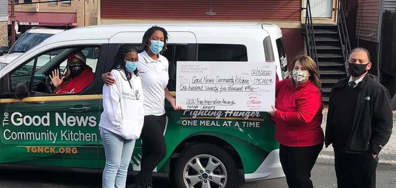 A group of people stand in front of a van labeled "The Good News Community Kitchen," holding an oversized check.