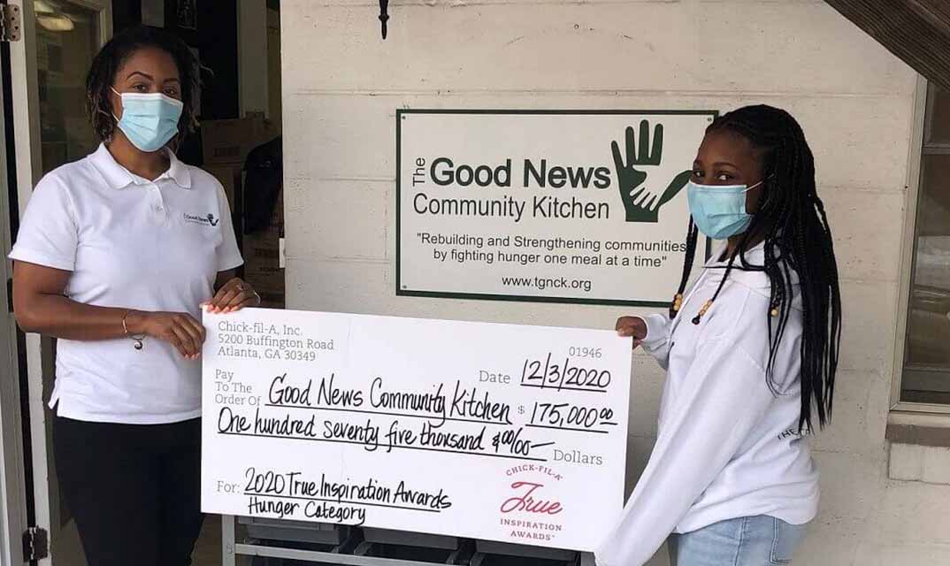 Two people stand in front of The Good News Community Kitchen, holding an oversized check.