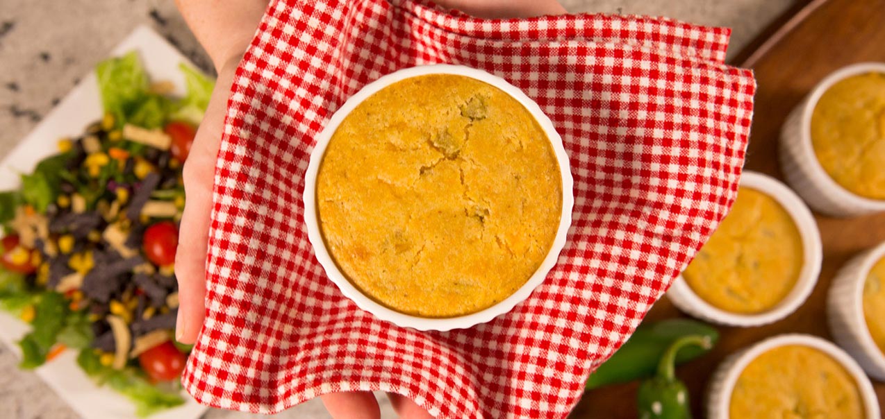 Cornbread pot pie in a white ramekin held with a red checkered cloth, with salad and more pot pies in the background.