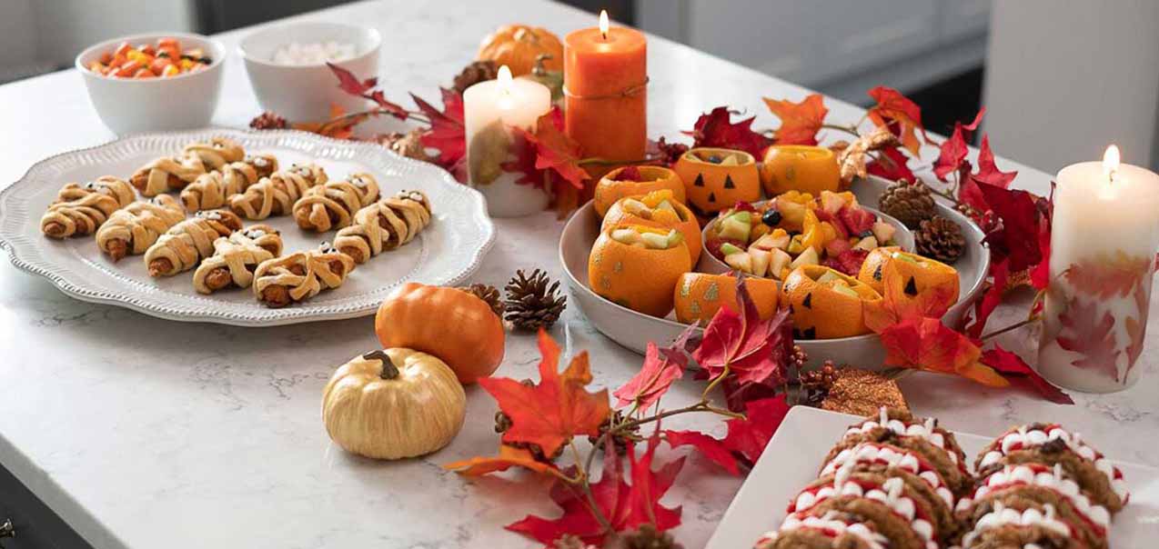 Halloween-themed table with mummy chicken wraps, jack-o'-lantern fruit cups, decorated cookies, candles, and autumn leaves.