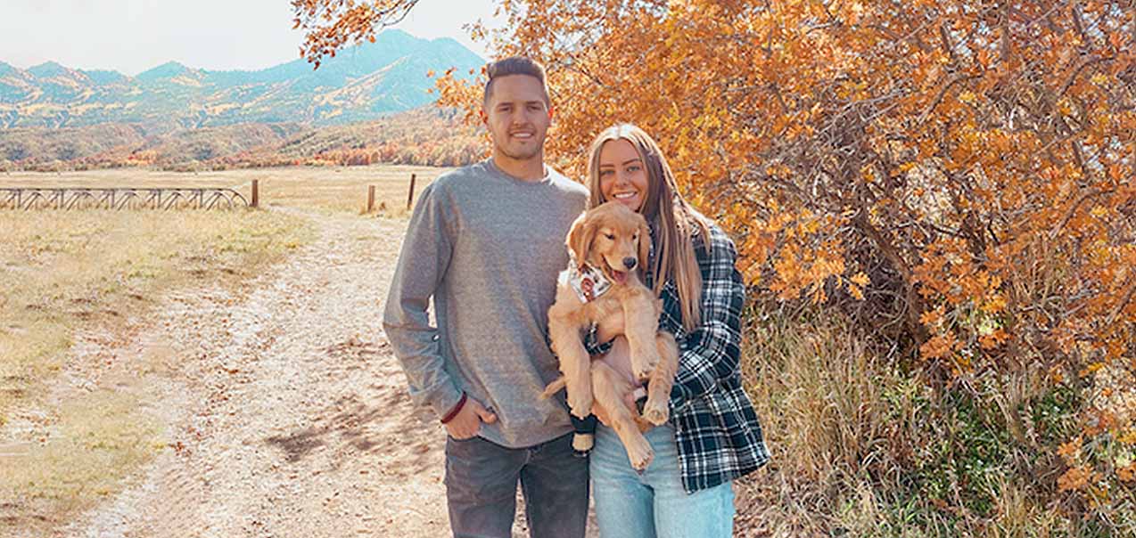 A young couple poses with their puppy against a fall background.