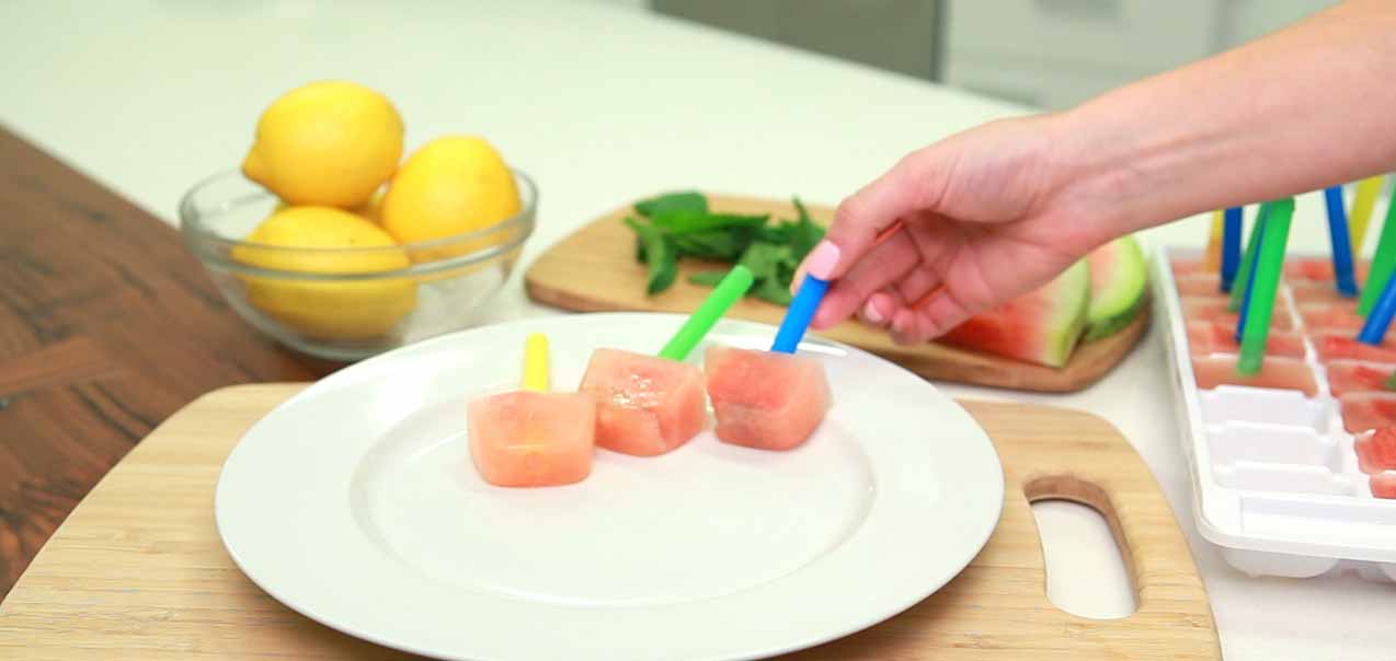 A hand places a pink popsicle with a blue stick on a white plate next to other popsicles, with lemons and mint in the background.