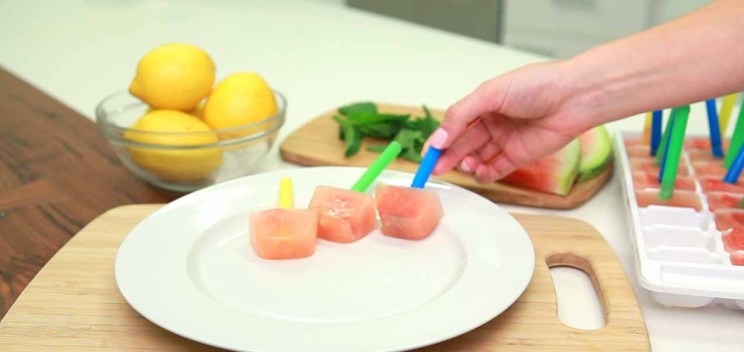 A hand places a pink popsicle with a blue stick on a white plate next to other popsicles, with lemons and mint in the background.