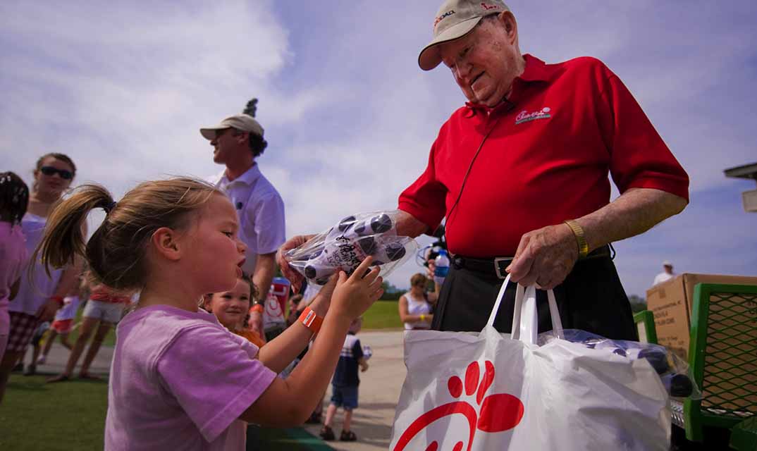 Truett Cathy handing a little girl a stuffed cow toy.