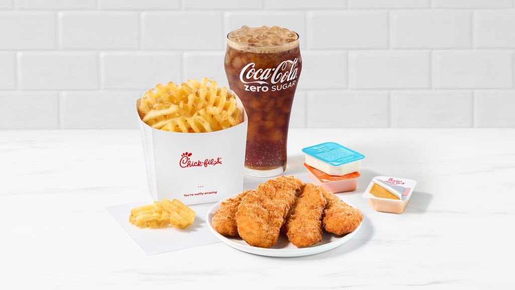 A plate of 4-count Chick-fil-A Chick-n-Strips® next to packets of Garden Herb Ranch Sauce, Zesty Buffalo Sauce and Chick-fil-A® Sauce with Waffle Fries and a Coca-Cola Zero on a white countertop with a white tiled background