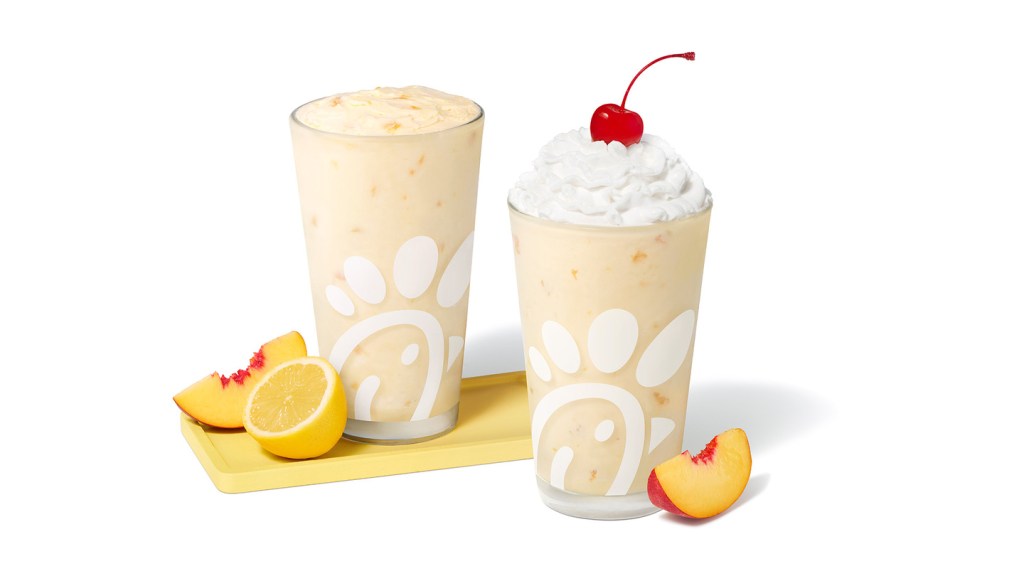 Peach Frosted Lemonade in a glass accompanied by a peach and a lemon slice and Peach milkshake in a glass topped with whipped cream and a cherry, accompanied by a fresh peach slice.