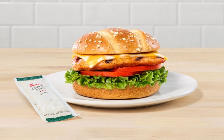 A boneless chicken breast on a toasted multigrain brioche bun, topped with Colby-Jack cheese, green leaf lettuce, and tomato slices. A small packet of Cilantro Lime Sauce sits beside the sandwich.