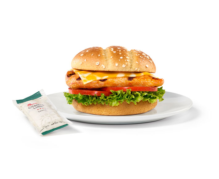 Grilled spicy chicken breast with peppers, served on a toasted multigrain brioche bun with Colby-Jack cheese, green leaf lettuce, and tomato, with a packet of Cilantro Lime sauce next to it.
