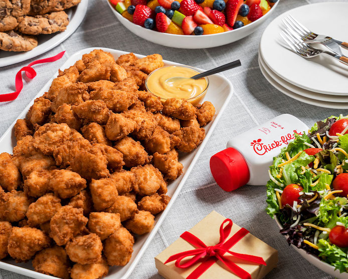 A festive table spread featuring a tray filled with Chick-fil-A Nuggets next to a bowl of salad, a bowl of fruit and a tray of cookies.