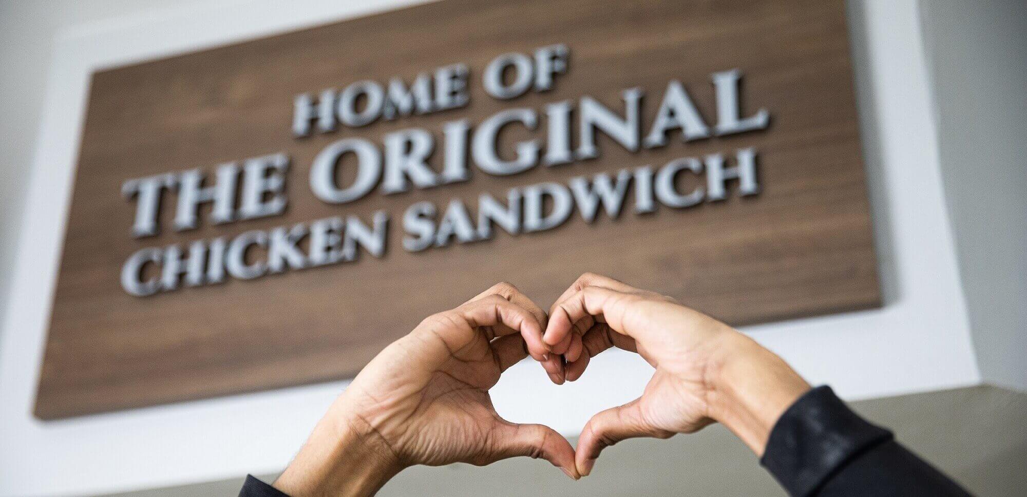 Hands forming a heart shape in front of a sign, with the text HOME OF THE ORIGINAL CHICKEN SANDWICH.