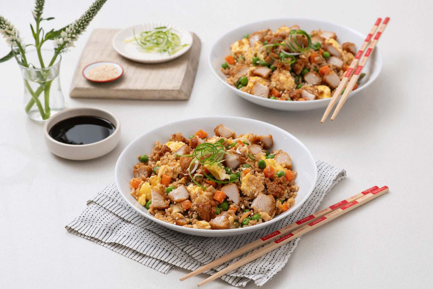 A bowl of fried rice with chicken, peas, carrots, and scrambled eggs, garnished with green onions, next to soy sauce and chopsticks.