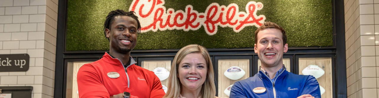 Three people standing in front of a Chick-fil-A logo and a wall with displayed footballs.
