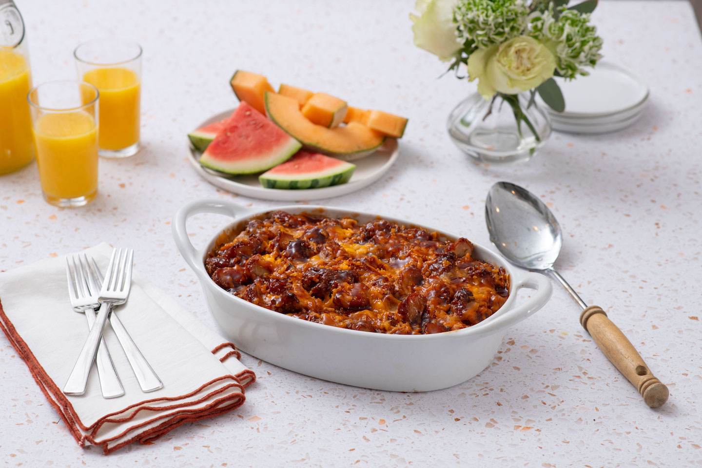 White casserole dish with baked casserole, forks and napkins, and a plate of watermelon slices.