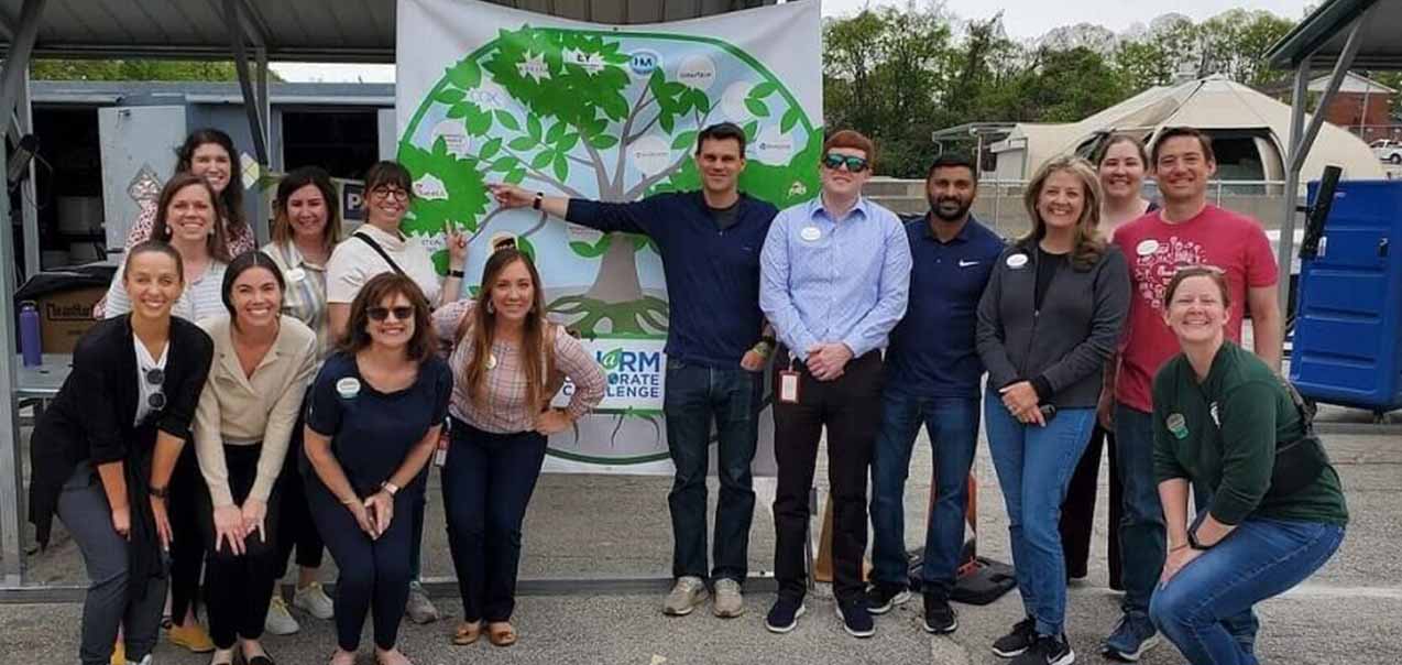 A group of thirteen people posing in front of a banner with a green tree graphic.