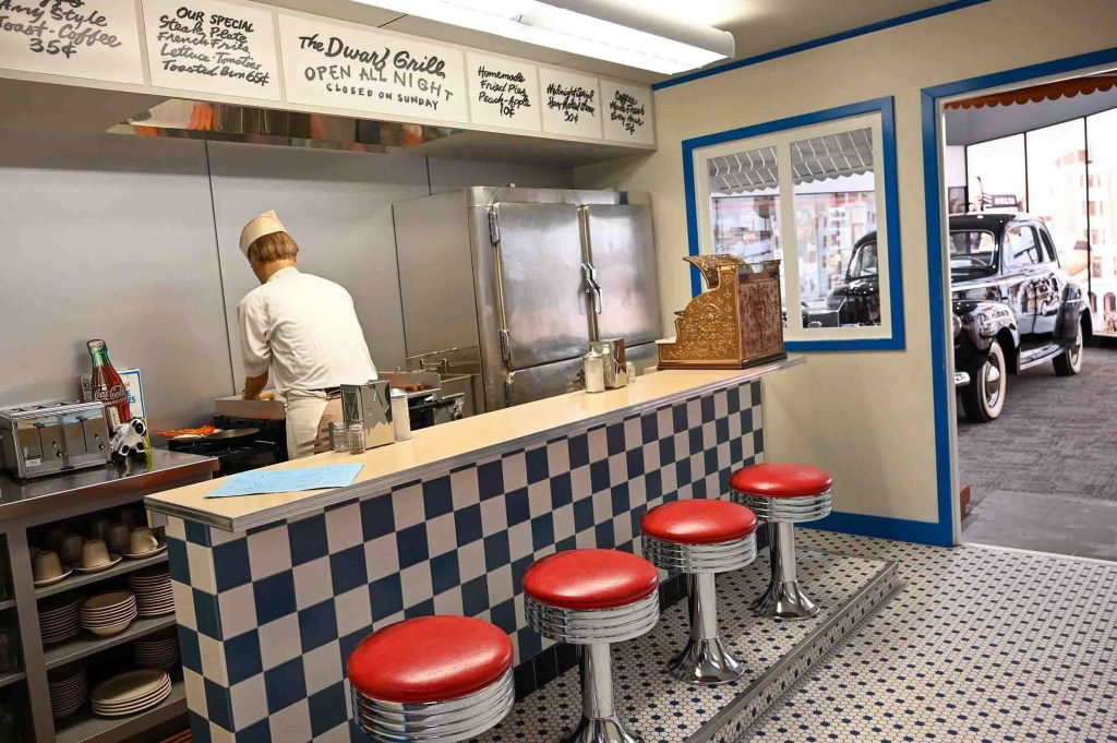 A retro diner with red bar stools, a cook in a white uniform, vintage decor, and a black vintage car in the background.