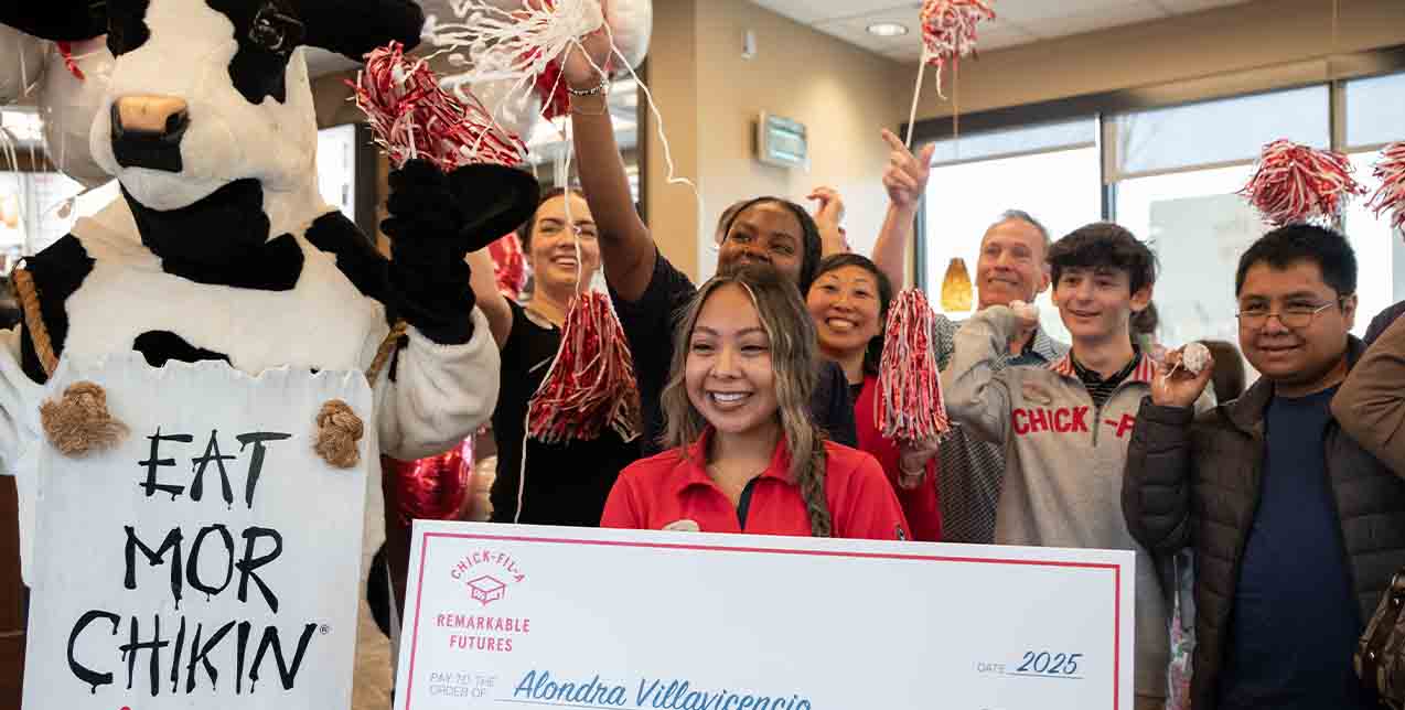 A team member standing together with a Chick-fil-A Cow & other team members, holding a check and smiling