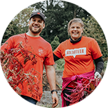 Two volunteers in orange shirts holding plants with reddish leaves outdoors.

