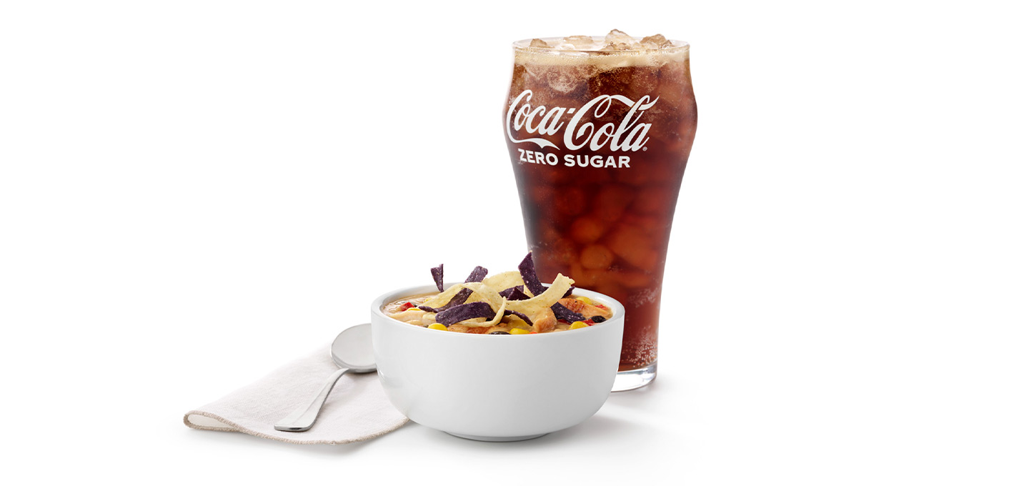 A bowl of Chicken Tortilla Soup next to a spoon on a napkin and a glass of Coca-Cola Zero on a white tile background
