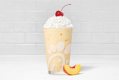 Peach milkshake in a glass topped with whipped cream and a cherry, accompanied by a fresh peach slice.
