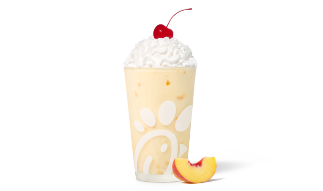 Peach milkshake in a glass topped with whipped cream and a cherry, accompanied by a fresh peach slice.