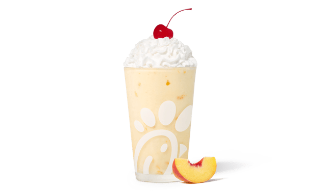 Peach milkshake in a glass topped with whipped cream and a cherry, accompanied by a fresh peach slice.