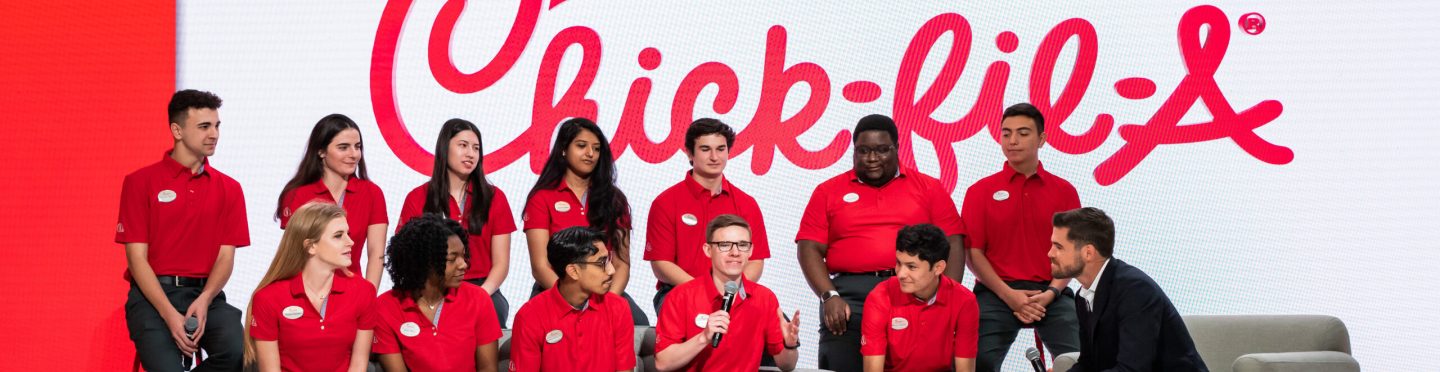 Group of Chick-fil-A team members in red uniforms seated on stage in front of a large Chick-fil-A logo, engaged in a discussion with a man in a suit.
