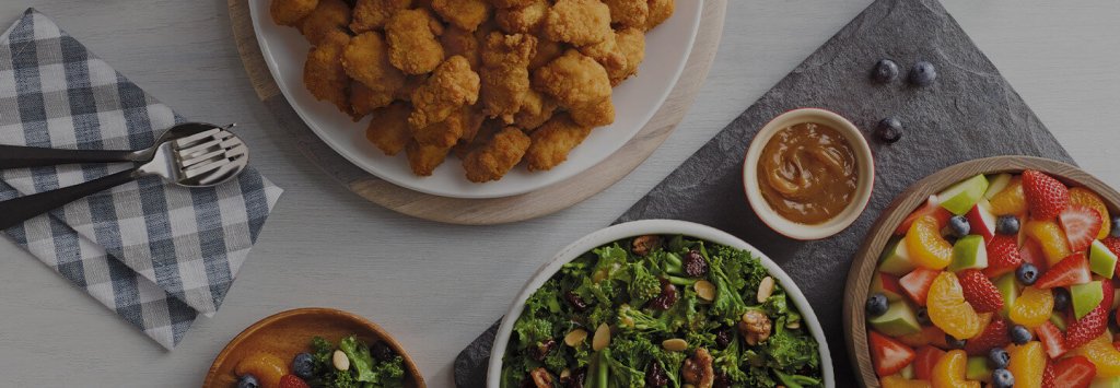 An array of catering items including chicken, a small bowl of sauce, a fruit salad, and a kale salad on a table.