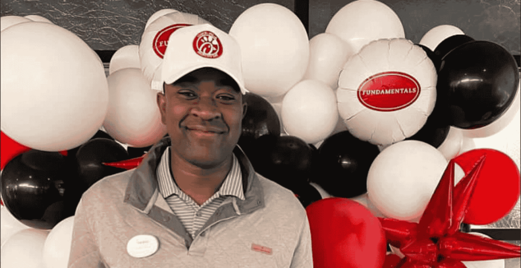 A person wearing a white cap and gray jacket stands in front of black, white, and red balloons. One balloon reads "FUNDAMENTALS."