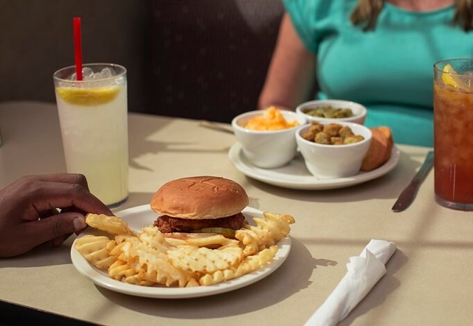 A plate of a Chicken Sandwich and Waffle Fries and a plate of okra, green beans and Mac & Cheese sitting on a table with drinks in front of customers