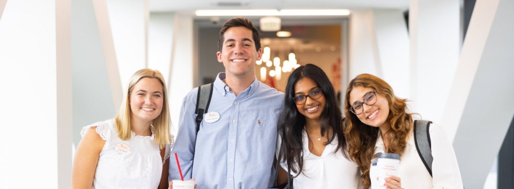 Four young adults standing indoors, smiling, with one holding a Chick-fil-A drink cup.