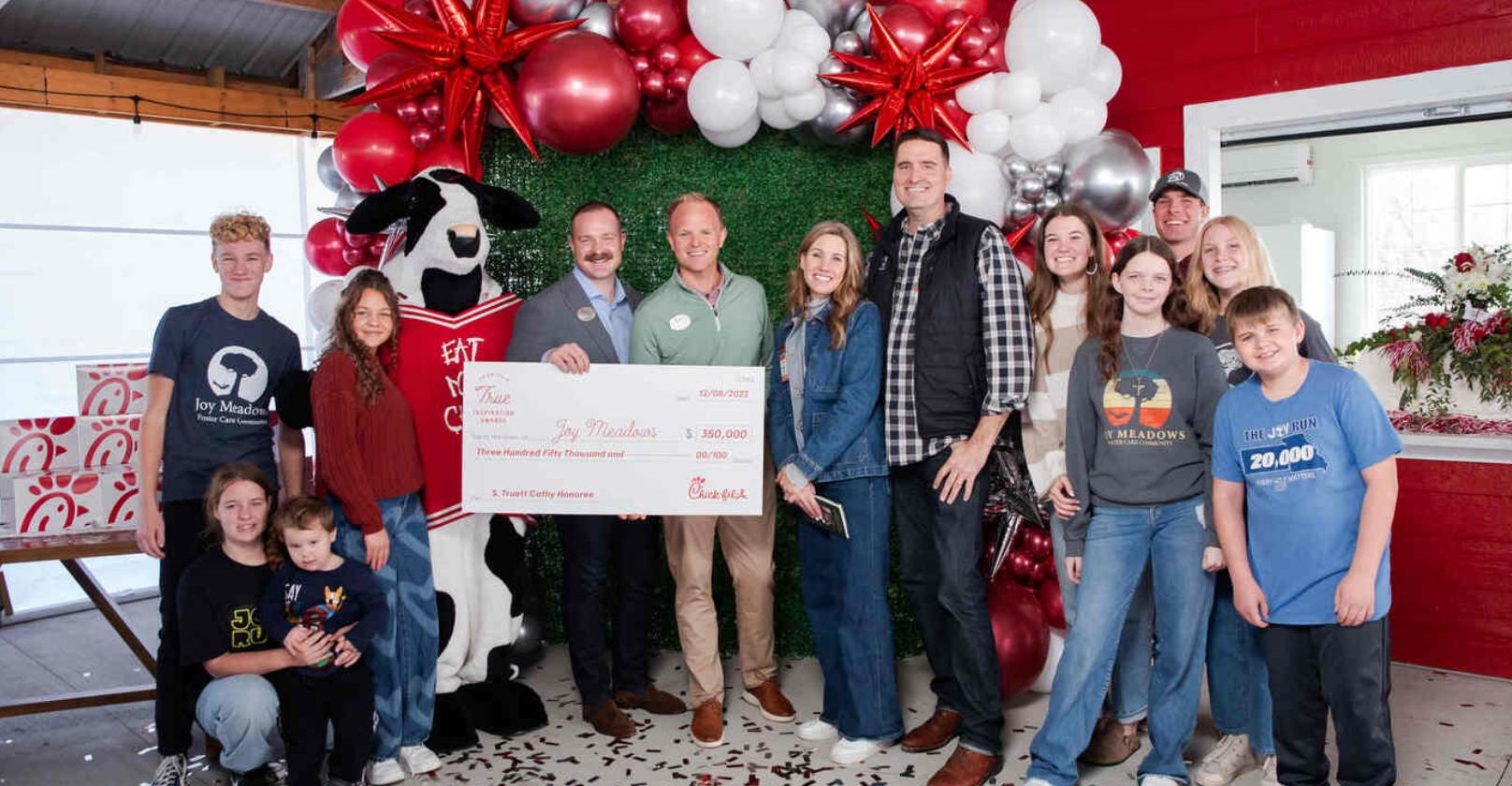 A group of people with a cow mascot holding a large check, surrounded by red, white, and silver balloon decorations.