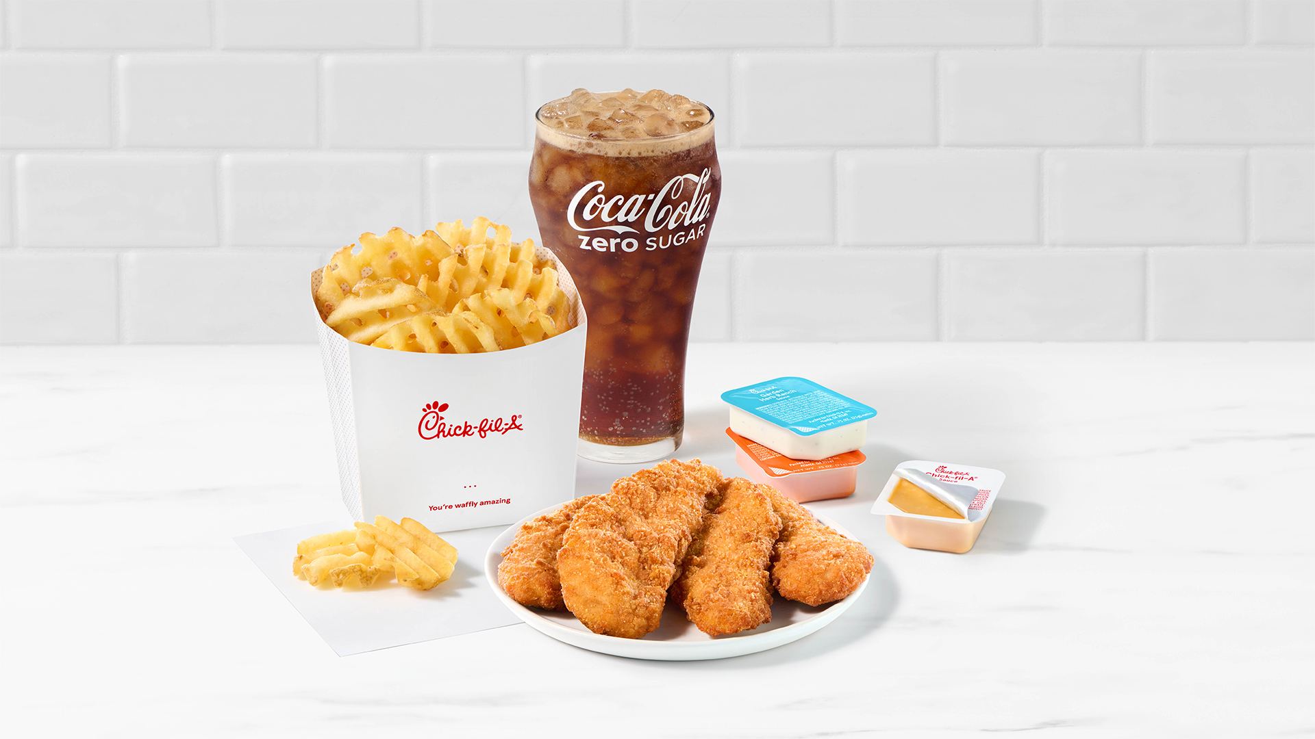 A plate of 4-count Chick-fil-A Chick-n-Strips® next to packets of Garden Herb Ranch Sauce, Zesty Buffalo Sauce and Chick-fil-A® Sauce with Waffle Fries and a Coca-Cola Zero on a white countertop with a white tiled background