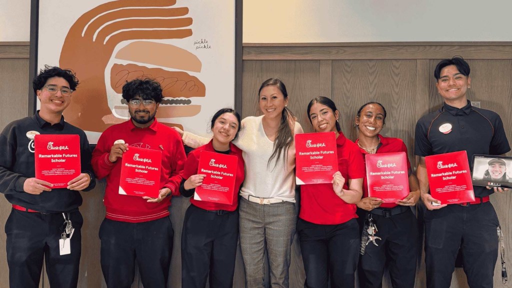 Six people, five in Chick-fil-A uniforms, holding "Remarkable Futures Scholar" certificates, standing indoors with a sandwich graphic in the background.