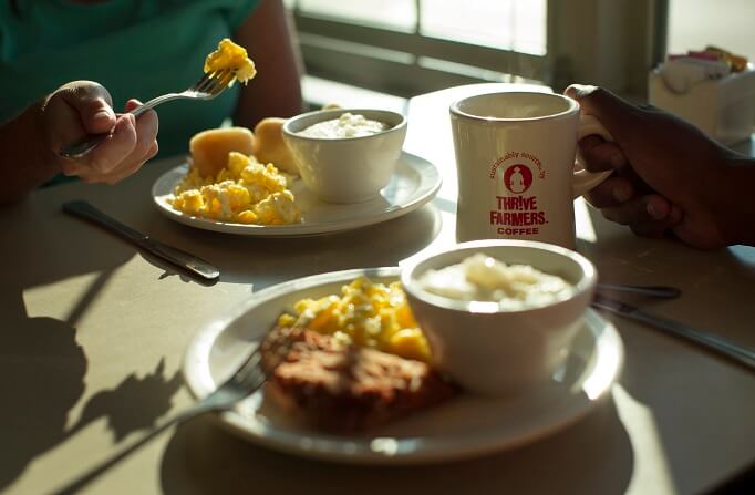 A table with breakfast plates and a “Thrive Farmers Coffee” mug, showcasing grits and scrambled eggs.