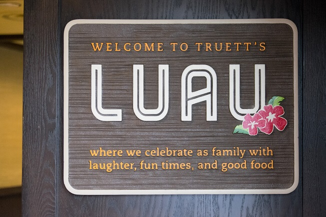 Sign reading "Welcome to Truett's Luau where we celebrate as family with laughter, fun times, and good food." with decorative hibiscus flowers.

