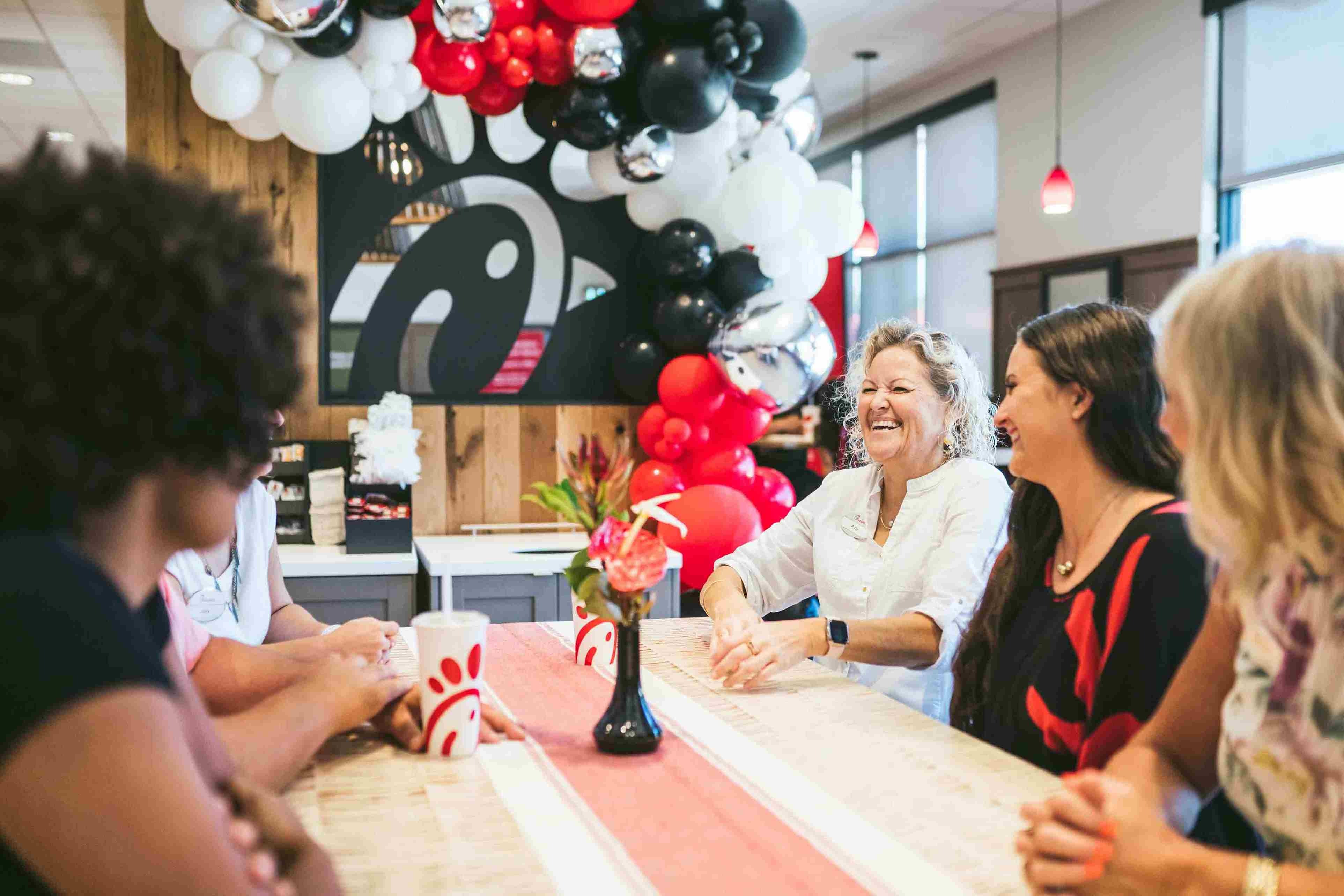 A group of people enjoying a lively conversation at a table decorated with an arch of red, black, and white balloons.