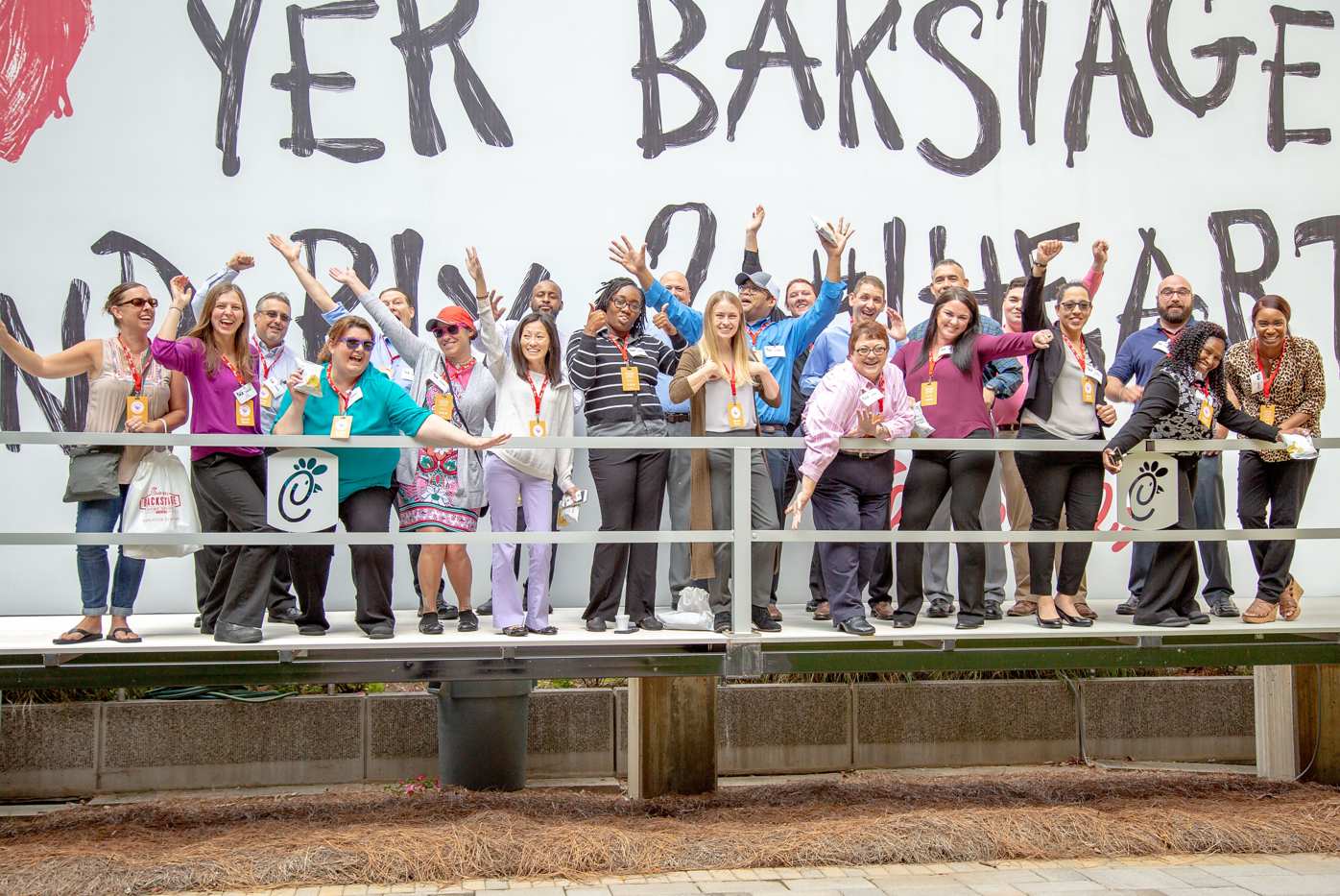 A group of people on a platform in front of a wall with large black letters, posing energetically and smiling.