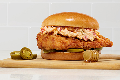 Honey Pepper Pimento Chicken Sandwich with chicken, pimento cheese, pickles, on a bun, with jalapeños and honey on a board.