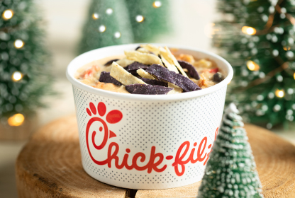 A cup of Chicken Tortilla Soup in a branded cup on a wooden surface with artificial evergreen trees in the background.