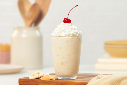 Banana Pudding Milkshake in a glass on a wooden board surrounded by ingredients & utensils.