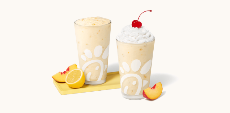 Peach Frosted Lemonade in a glass accompanied by a peach and a lemon slice and Peach milkshake in a glass topped with whipped cream and a cherry, accompanied by a fresh peach slice.