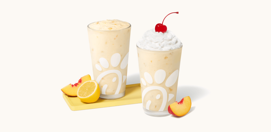 Peach Frosted Lemonade in a glass accompanied by a peach and a lemon slice and Peach milkshake in a glass topped with whipped cream and a cherry, accompanied by a fresh peach slice.