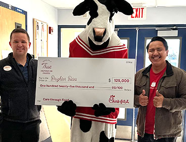 A Chick-fil-A Cow holding a large check and standing with two people from Brighter Bites