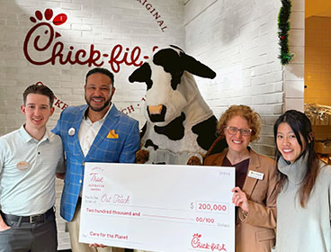 People from the Out Teach organization standing together smiling with a Chick-fil-A Cow and holding a large check
