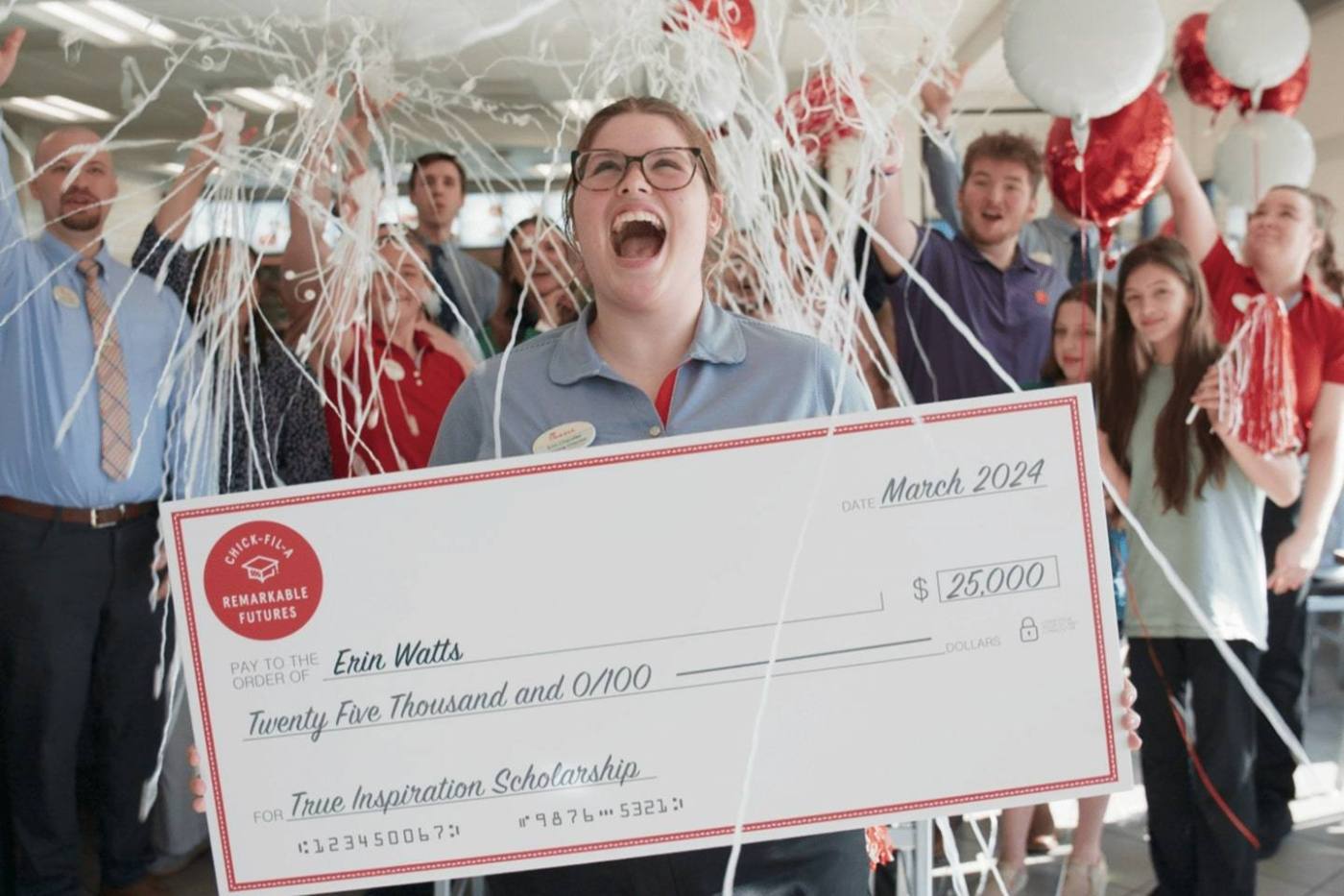 A person celebrates with a group, holding a large ceremonial check surrounded by balloons and streamers.