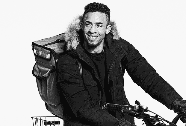 Black-and-white GIF of a smiling food delivery worker riding a bicycle, wearing a winter coat and delivery backpack.