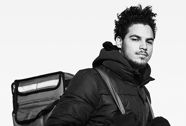 Black-and-white GIF of a smiling food delivery worker riding a bicycle, wearing a winter coat and delivery backpack.