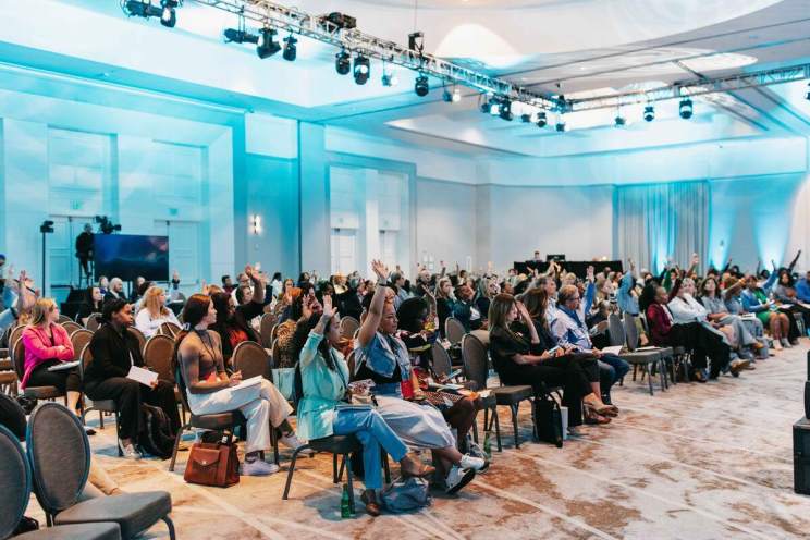 2025 Impact Accelerator Conference attendees seated and raising their hands in a large room with blue lighting.
