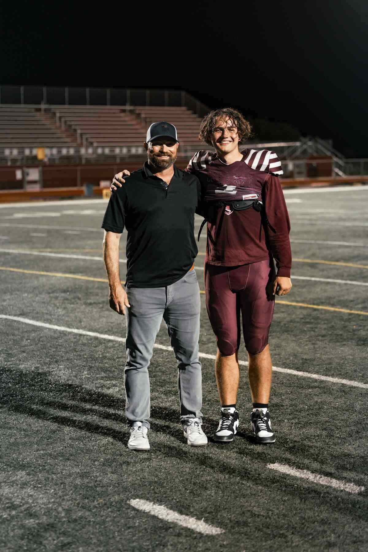 Two people smiling on a football field at night, one in a black polo and cap, the other in a maroon football uniform.